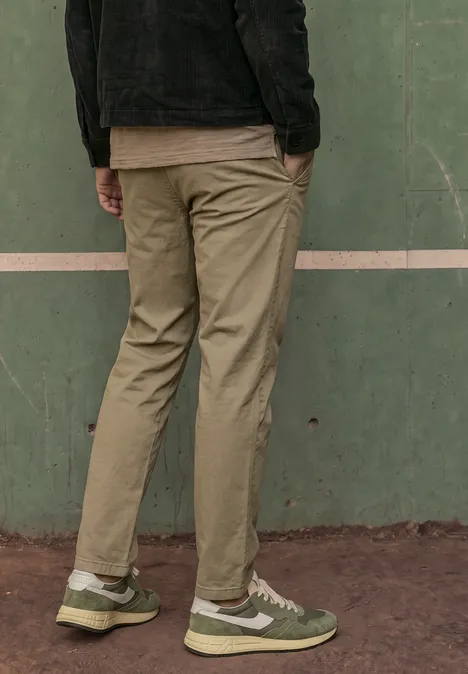 Person steht vor grüner Wand, trägt olivgrüne Hose und grüne Sneaker.