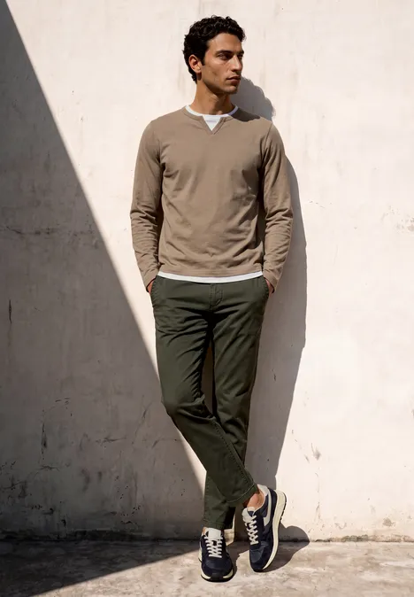Ein Mann lehnt lässig an eine helle Wand, trägt olivgrüne Hose, braunen Pullover und Turnschuhe im Schatten.