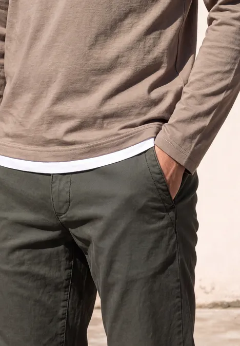 Person in hellbraunem Langarmshirt über weißem T-Shirt und dunkelgrüner Hose, Hand in Tasche