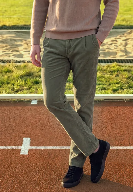 Eine Person in rosafarbenem Pullover, grünen Cordhosen und schwarzen Schuhen steht lässig auf einer Laufbahn.