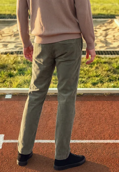 Eine Person von hinten in beiger Jacke und olivgrünen Hosen steht auf einer Tartanbahn neben einem Sandkasten.