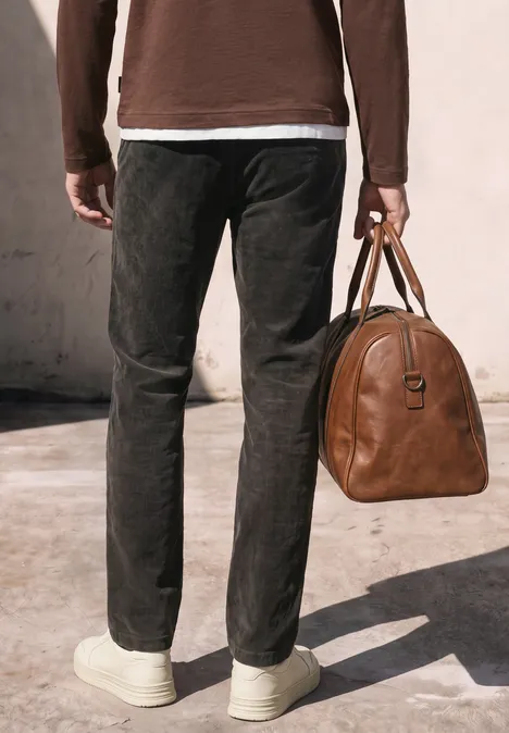 Mann trägt schwarze Cordhose, weiße Sneaker, hält braune Ledertasche, steht vor hellem Hintergrund.