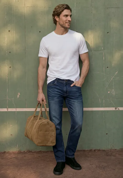 Straight Leg Jeans im Regular Fit indigo blue wash Ein Mann in weißem T-Shirt und blauen Jeans steht vor einer grünen Wand und hält eine braune Tasche.