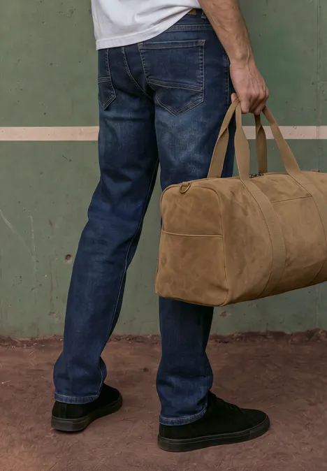 Straight Leg Jeans im Regular Fit indigo blue wash Mann trägt blaue Jeans und schwarze Schuhe, hält eine beige Reisetasche vor einer grünen Wand.
