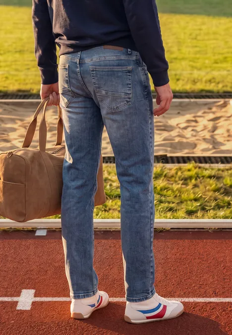 Slim Leg-jeans met Regular Fit soft greencast random wash Man in spijkerbroek en sneakers met reistas staat op baan naast zandbak.
