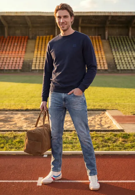 Slim Leg-jeans met Regular Fit soft greencast random wash Een man staat op de atletiekbaan voor de verspringzandbak, houdt een tas vast, draagt jeans en trui.