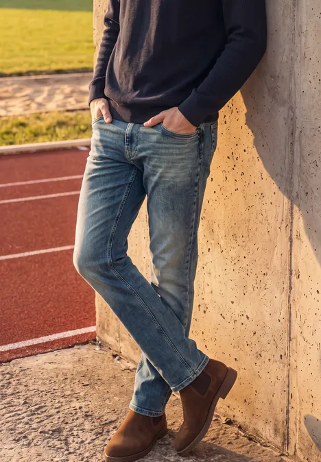 Straight Leg Jeans im Regular Fit soft greencast random wash Ein Mann in Jeans und Pullover lehnt entspannt an einer Betonwand neben einer Laufbahn.