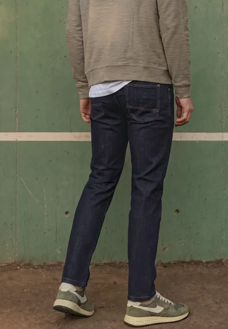 Person mit dem Rücken zur Kamera, trägt dunkle Jeans und Sneaker vor grüner Wand.