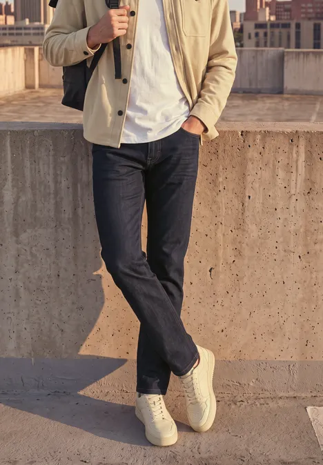 Un homme en jean foncé, veste beige et baskets blanches, debout devant un mur en béton avec un décor urbain.