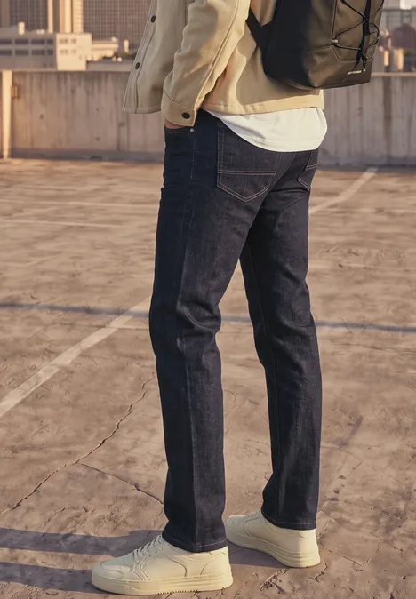Personne en jean sombre et baskets blanches, portant un sac à dos sur un toit en béton avec vue urbaine.