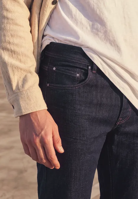 Gros plan latéral d’une personne au bord de la plage en jean foncé, t-shirt blanc et veste beige.