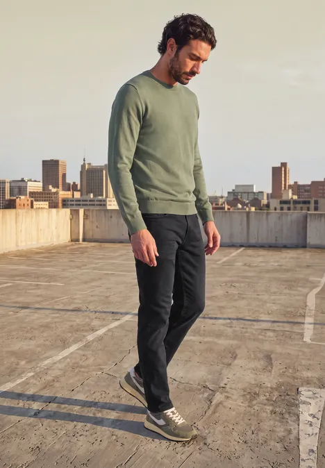 Ein Mann in grünem Pullover und dunkler Hose geht auf einem Parkdeckdach mit Stadtkulisse.