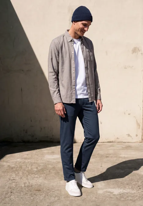 Mann mit Mütze, offenes Hemd über T-Shirt, schmaler Hose und weißen Sneakern steht entspannt in Sonnenlicht.