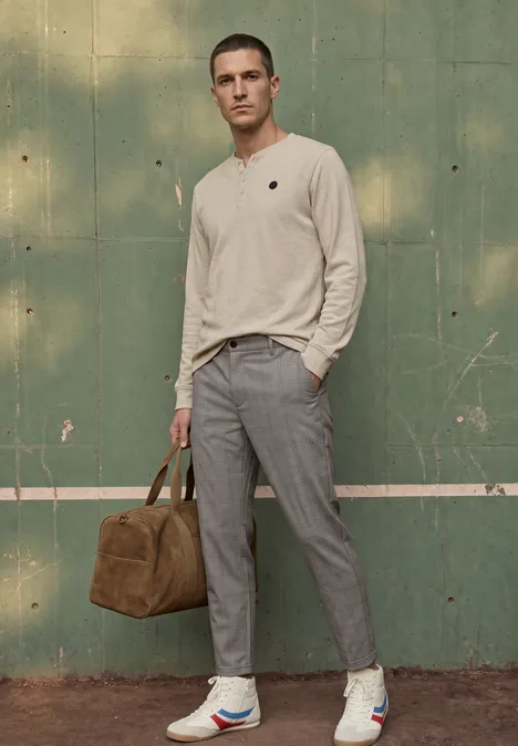 Ein Mann in beigem Pullover, grauen Hosen und Sneakers steht vor grüner Wand und trägt eine braune Reisetasche.