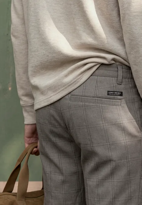 Rückenansicht einer Person in beigem Pullover und grauen Karohosen, die eine braune Tasche hält.