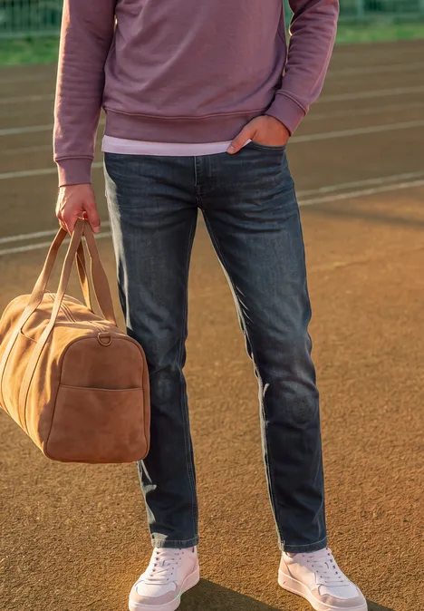 Jean Slim Leg à coupe classique dark blue coloured back Personne en jean, baskets blanches et sweat rose tient un sac de voyage marron sur une piste.