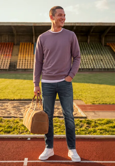 Jean Slim Leg à coupe classique dark blue coloured back Un homme en sweat violet, jean et baskets blanches porte un sac de sport sur la piste du stade.