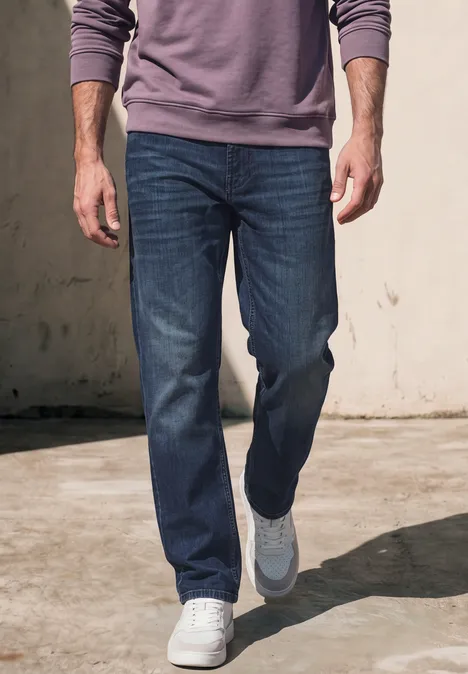 Man in blauwe jeans en witte sneakers loopt voor een lichte muur.