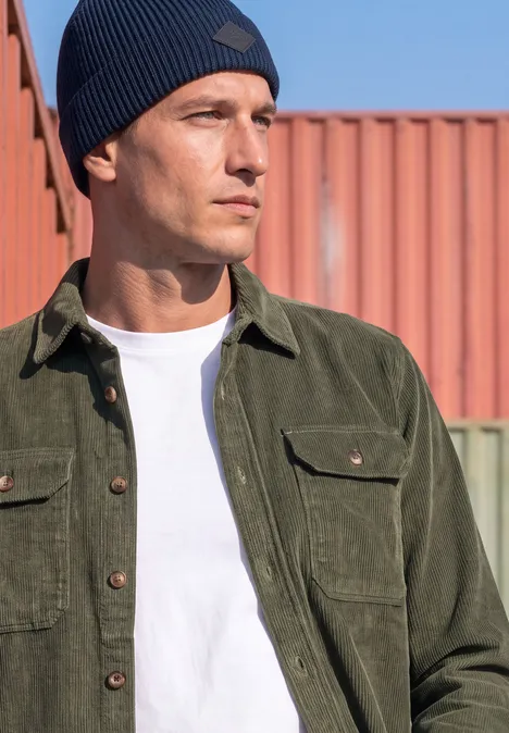 Un homme en t-shirt blanc, veste en velours côtelé verte et bonnet bleu regarde sur le côté en milieu urbain.