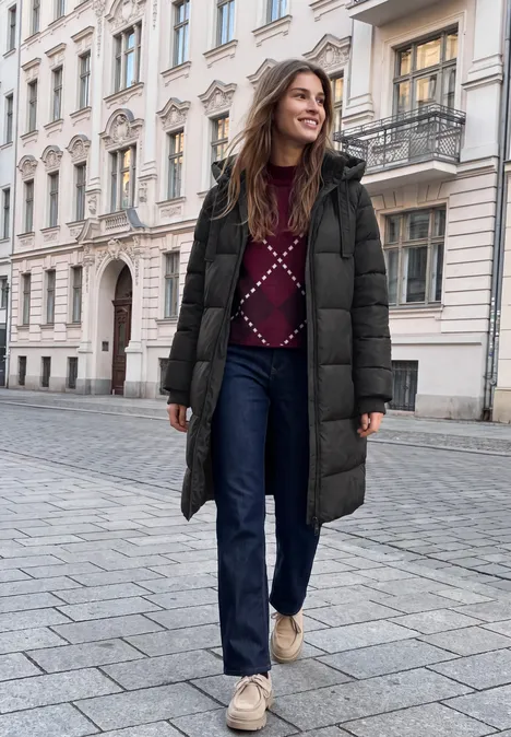 Eine Frau geht lächelnd in langer Daunenjacke, Pulli und Jeans eine städtische Straße entlang.