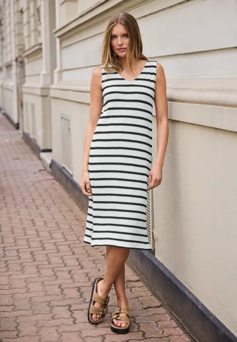 Frau in gestreiftem Midikleid und Sandalen lehnt an einer hellen Hauswand auf gepflastertem Gehweg.