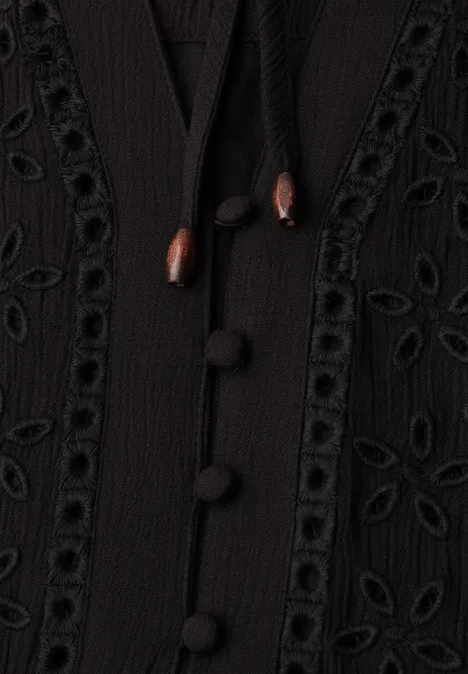 Close-up van een zwart kledingstuk met stofknopen, houten koordjes en fijne open borduursels.