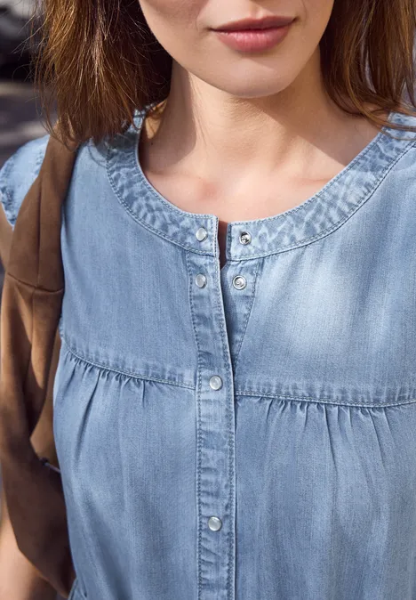 Teilaufnahme einer Frau in hellem Denim-Oberteil, sichtbar Hals, Brustbereich und brauner Taschenriemen.