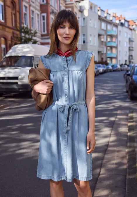 Eine Frau in ärmellosem Jeanskleid hält eine Clutch und geht eine sonnige Straße entlang.