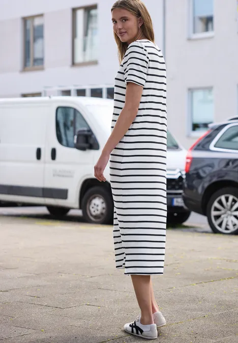 Vrouw in witte, zwart gestreepte midijurk en witte sandalen staat op stoep voor geparkeerde auto’s.