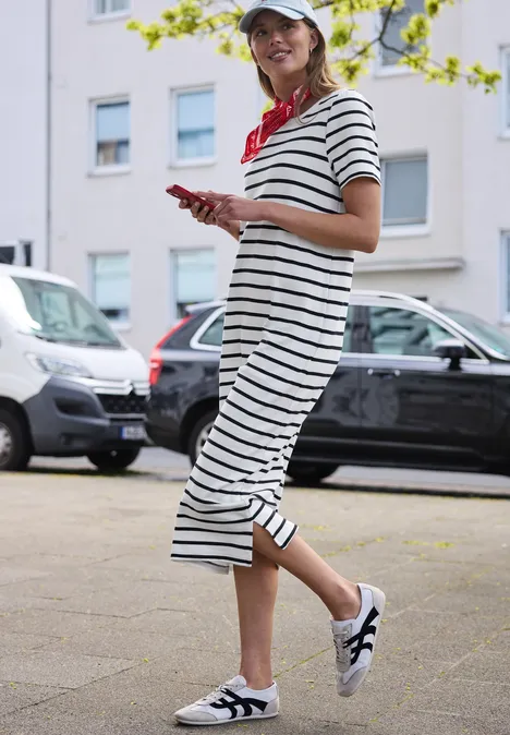 Een vrouw in gestreepte midijurk en sneakers loopt met haar smartphone op het trottoir.