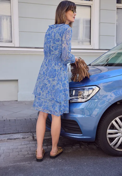 Een vrouw in een blauw gebloemde jurk staat met haar tas naast een kleine blauwe auto op straat.