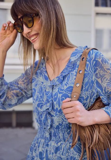 Vrouw draagt blauw, gemêleerd topje met taille-inzet en schoudertas, staat voor gevel.