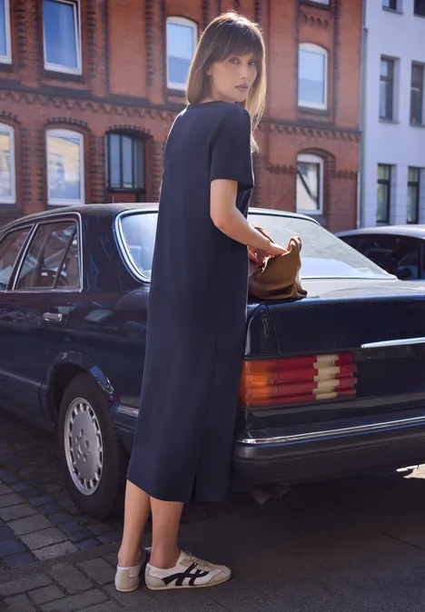 Een vrouw in een donkerblauwe midi-jurk en witte sneakers staat bij een zwarte auto en houdt een bruine tas vast.