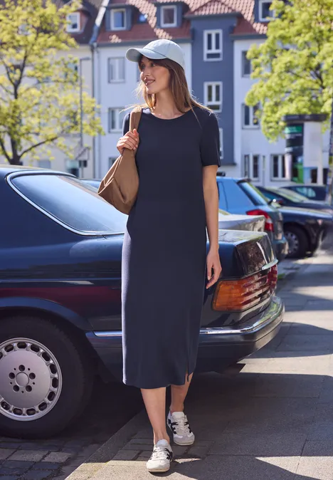 Vrouw in zwarte midijurk naast zwarte auto op een zonnige stadsstraat met moderne panden.