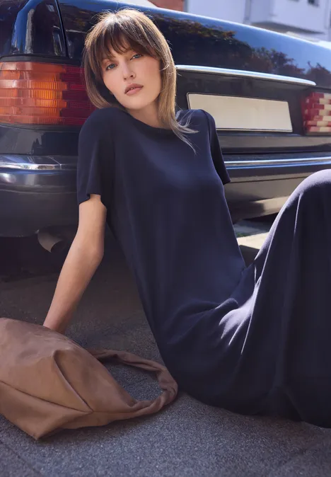 Een vrouw zit op de grond naast een geparkeerde auto, draagt een lange donkere jurk en heeft een tas naast zich.