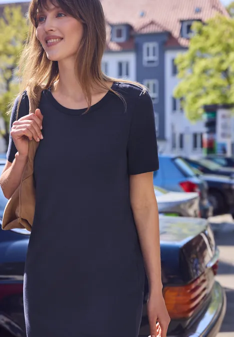 Vrouw in zwart T-shirtjurkje met schoudertas, lopend door een zonnige stadsstraat.
