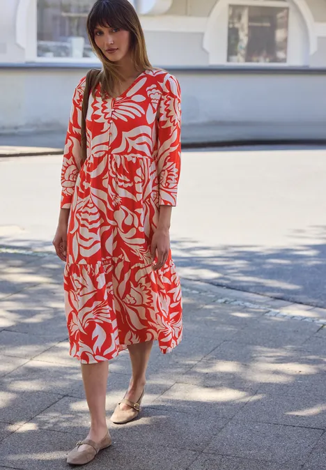 Vrouw in rode midi-jurk met wit patroon en sandalen staat op het trottoir voor een gebouw.