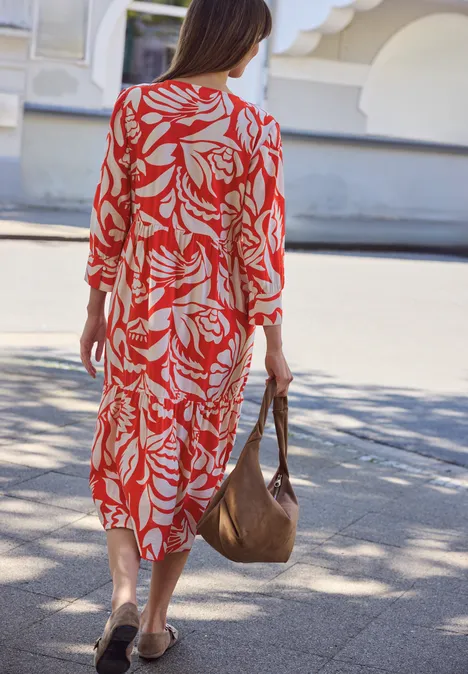 Vrouw in rode jurk met wit bladpatroon, met een bruine tas, loopt over een zonnig trottoir.