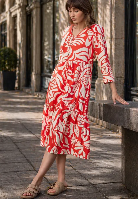 Eine Frau in rotem weißem Blumenmuster-Midikleid steht draußen an einer Steinbank.