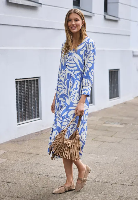 Frau in blau-weiß floralem Midikleid, mit Basttasche und Espadrilles vor hellem Haus