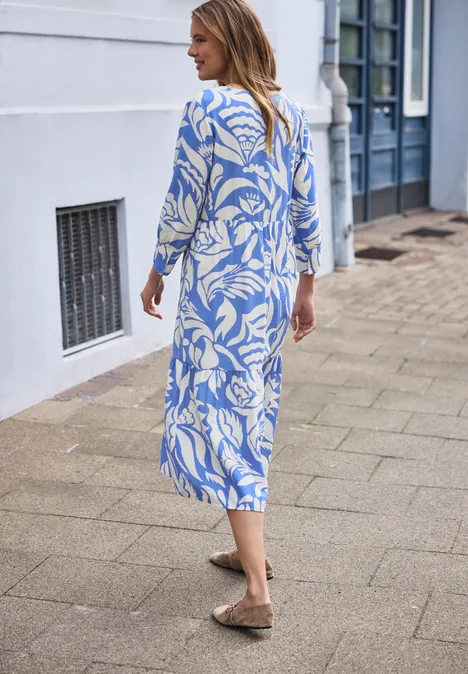 Eine Frau spaziert auf dem Bürgersteig in einem blau-weißen, floralen Maxikleid und braunen Slippern.