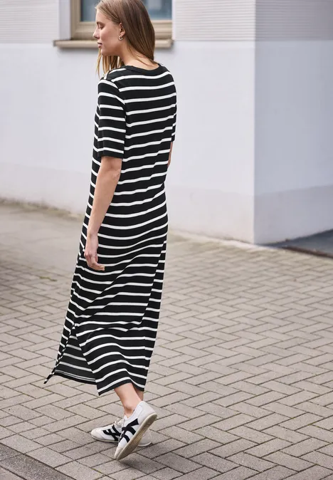 Een vrouw in een zwart-wit gestreepte maxijurk en sneakers loopt over een geplaveid pad.