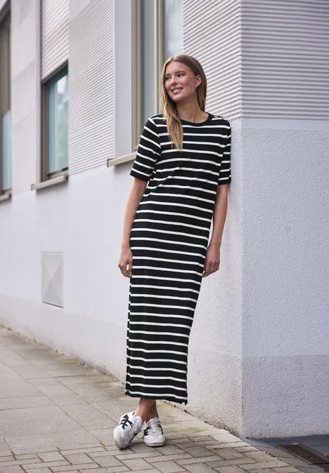 Vrouw in lange zwart-wit gestreepte jurk en witte sneakers, staand bij een gevel.