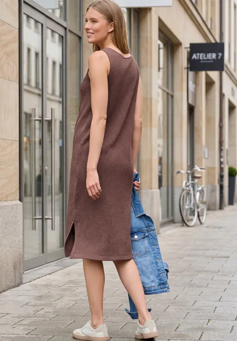 Eine Frau in braunem ärmellosem Kleid geht mit einer Jeansjacke in der Hand eine Straße entlang.