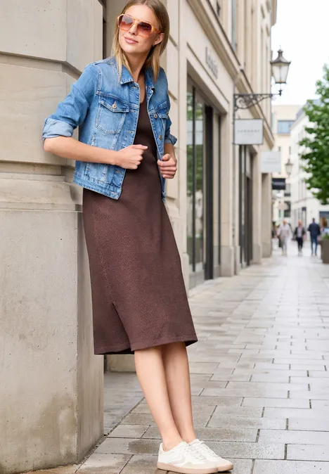 Eine Frau lehnt in lässiger Jeansjacke und braunem Kleid an einer Häuserwand in einer Fußgängerzone.