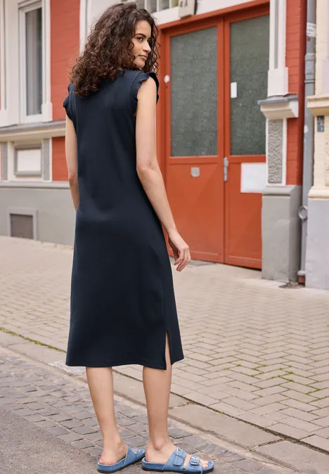 Een vrouw in een mouwloze zwarte jurk en blauwe sandalen staat voor een rode deur.