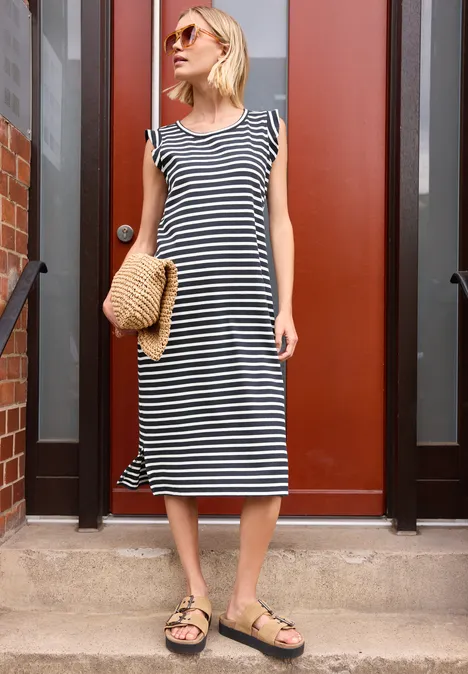 Eine Frau in gestreiftem Midi-Kleid trägt Birkenstock-Sandalen und hält einen Strohhut vor einer roten Tür.