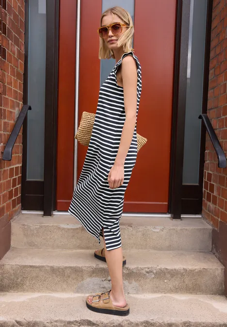 Frau in gestreiftem Midi-Kleid und Sandalen hält einen Strohtasche auf einer Treppe.