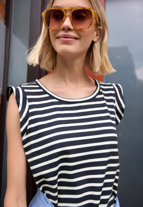 Eine Frau mit kurzem blondem Haar, Sonnenbrille und schwarz-weiß gestreiftem Top vor einem Gebäude.