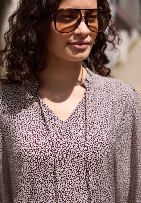 Knielanges Kleid mit V-Neck und Print vanilla sand Frau mit lockigem Haar trägt eine helle Bluse mit kleinem Blumenmuster und V-Ausschnitt.